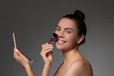 Smiling woman with mirror applying makeup on grey background