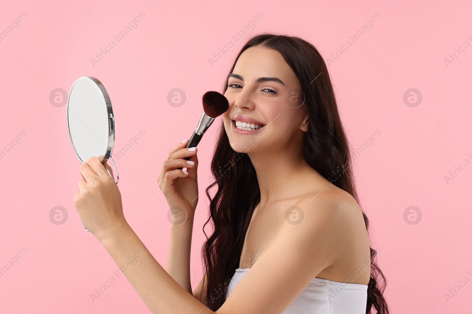 Photo of Smiling woman with mirror applying makeup on pink background