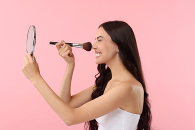 Smiling woman with mirror applying makeup on pink background