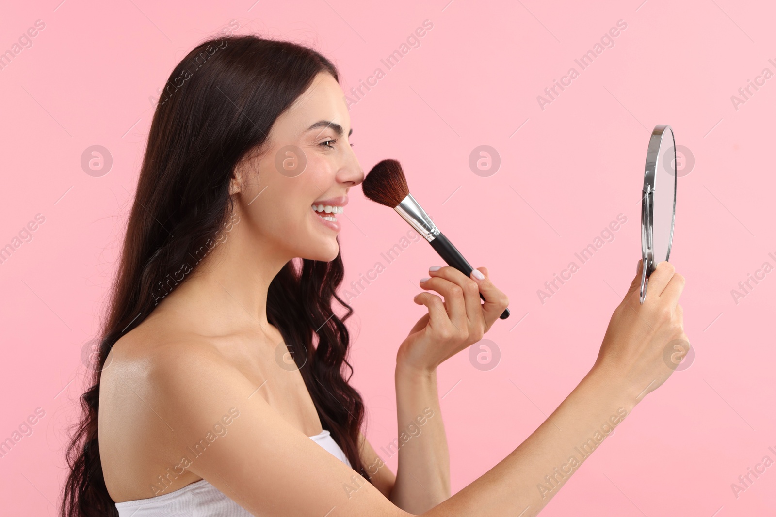 Photo of Smiling woman with mirror applying makeup on pink background