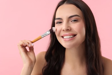 Smiling woman applying makeup with brush on pink background