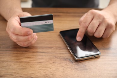 Photo of Man with credit card using smartphone at wooden table indoors, closeup