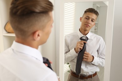 Photo of Handsome man adjusting necktie near mirror indoors