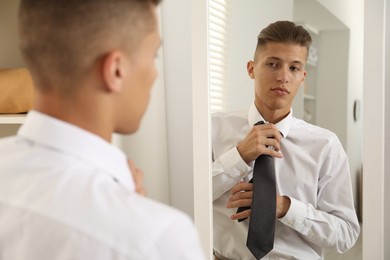 Photo of Handsome man adjusting necktie near mirror indoors