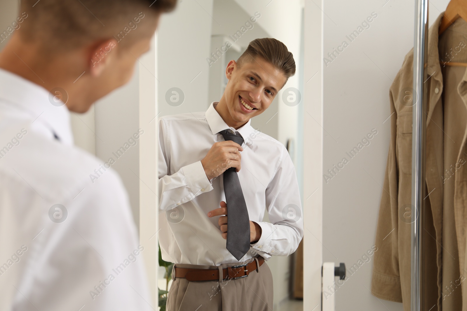 Photo of Handsome man adjusting necktie near mirror indoors