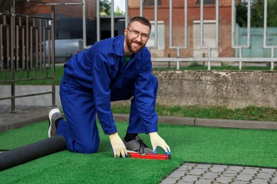 Photo of Happy young man in uniform installing artificial turf outdoors