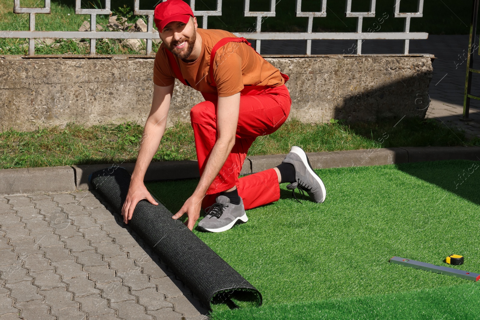 Photo of Happy young man in uniform installing artificial turf outdoors