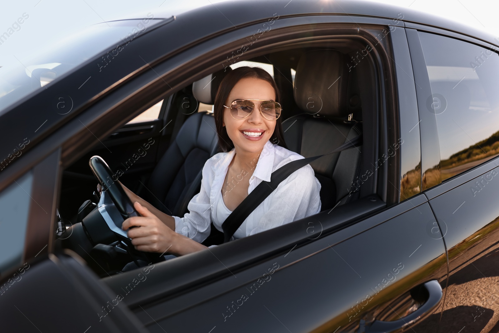 Photo of Smiling young woman in sunglasses with seatbelt driving car, view from outside