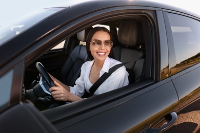 Smiling young woman in sunglasses with seatbelt driving car, view from outside