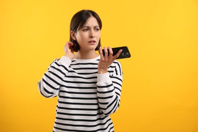 Beautiful woman recording voice message via smartphone on yellow background