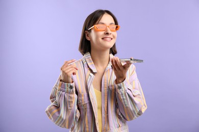 Beautiful woman in sunglasses recording voice message via smartphone on violet background