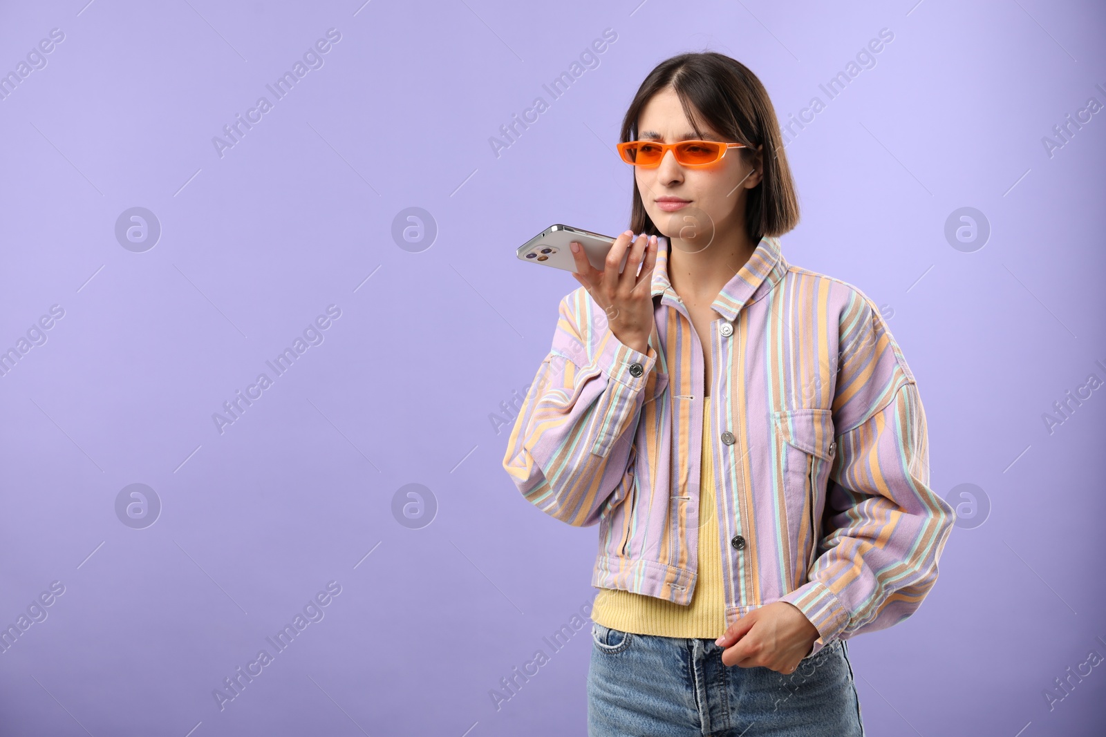 Photo of Beautiful woman in sunglasses recording voice message via smartphone on violet background, space for text