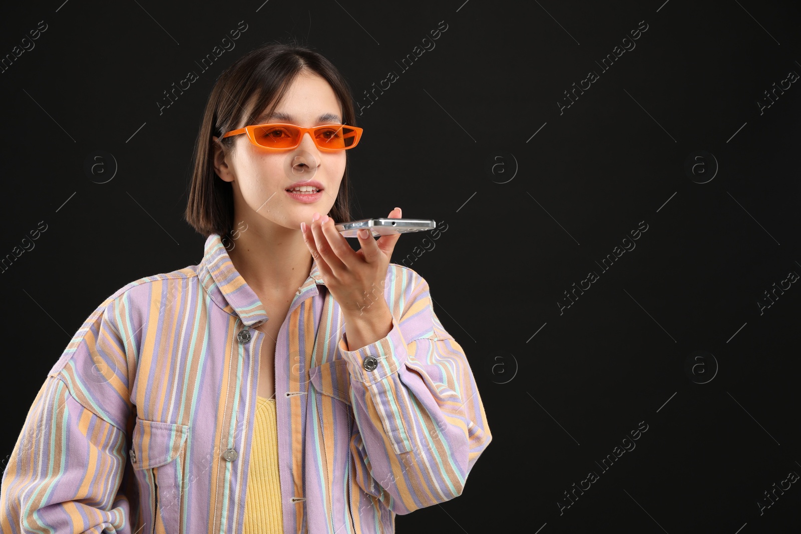 Photo of Beautiful woman in sunglasses recording voice message via smartphone on black background, space for text