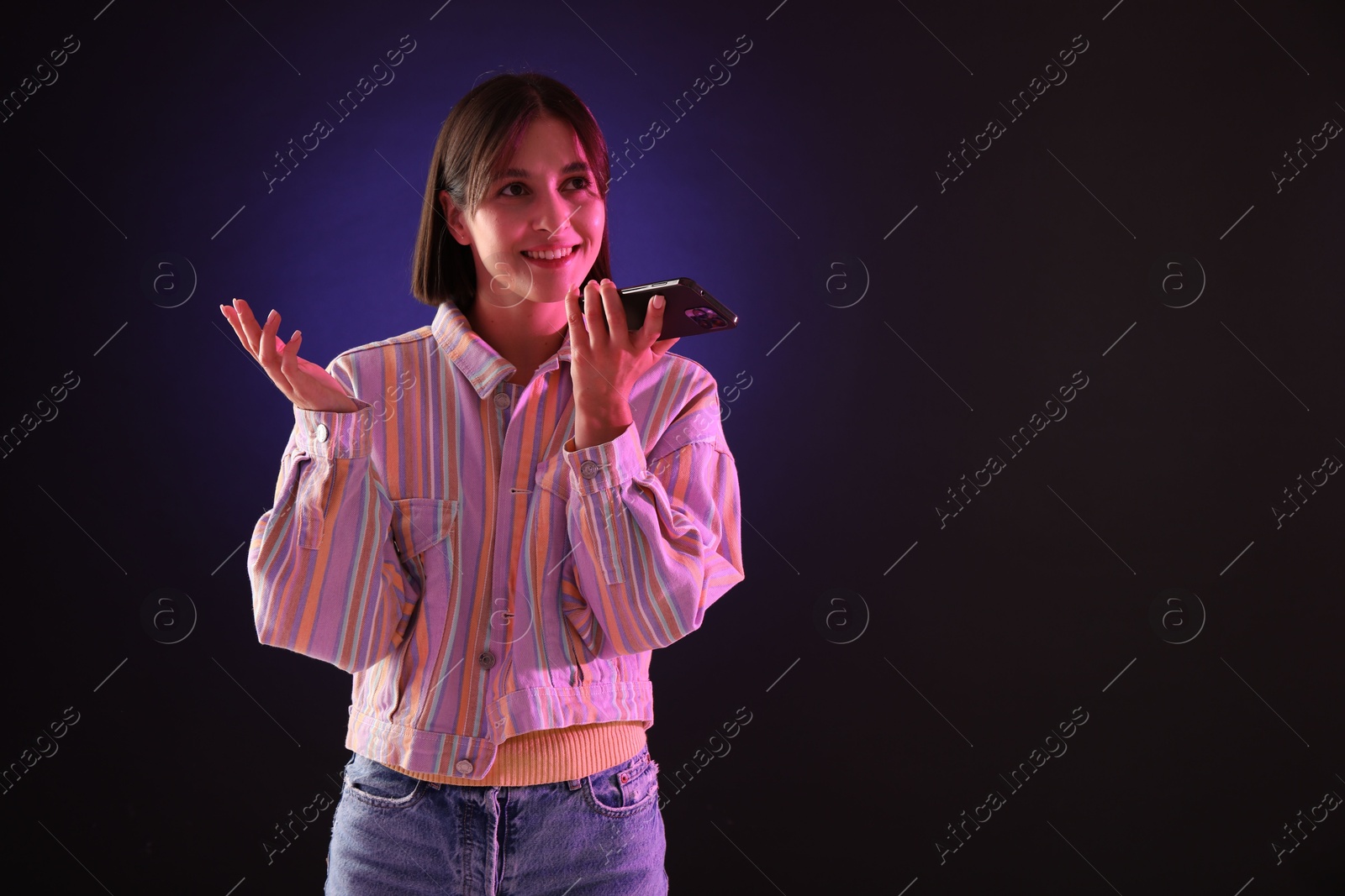 Photo of Beautiful woman recording voice message via smartphone on dark background with neon lights, space for text