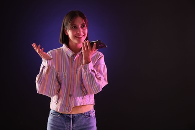 Beautiful woman recording voice message via smartphone on dark background with neon lights, space for text