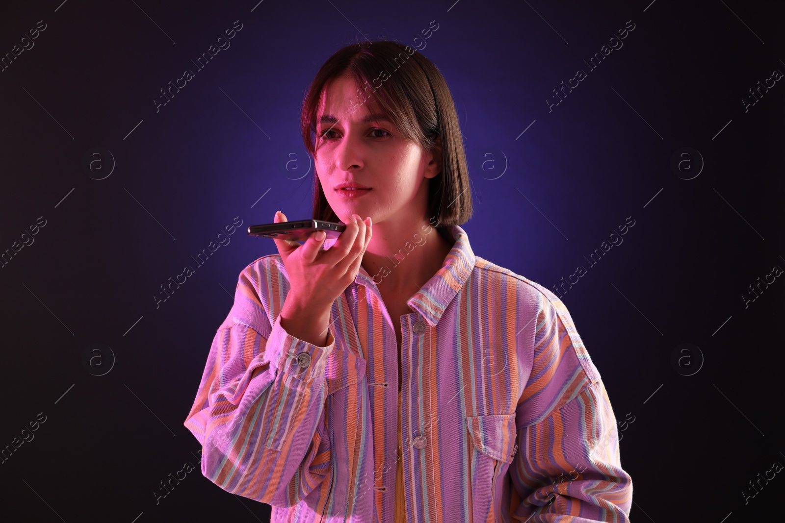 Photo of Beautiful woman recording voice message via smartphone on dark background with neon lights