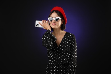 Stylish woman listening to voice message via smartphone on dark background