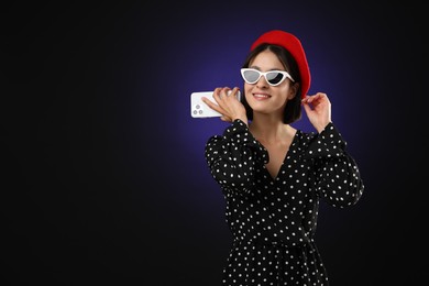 Stylish woman listening to voice message via smartphone on dark background, space for text