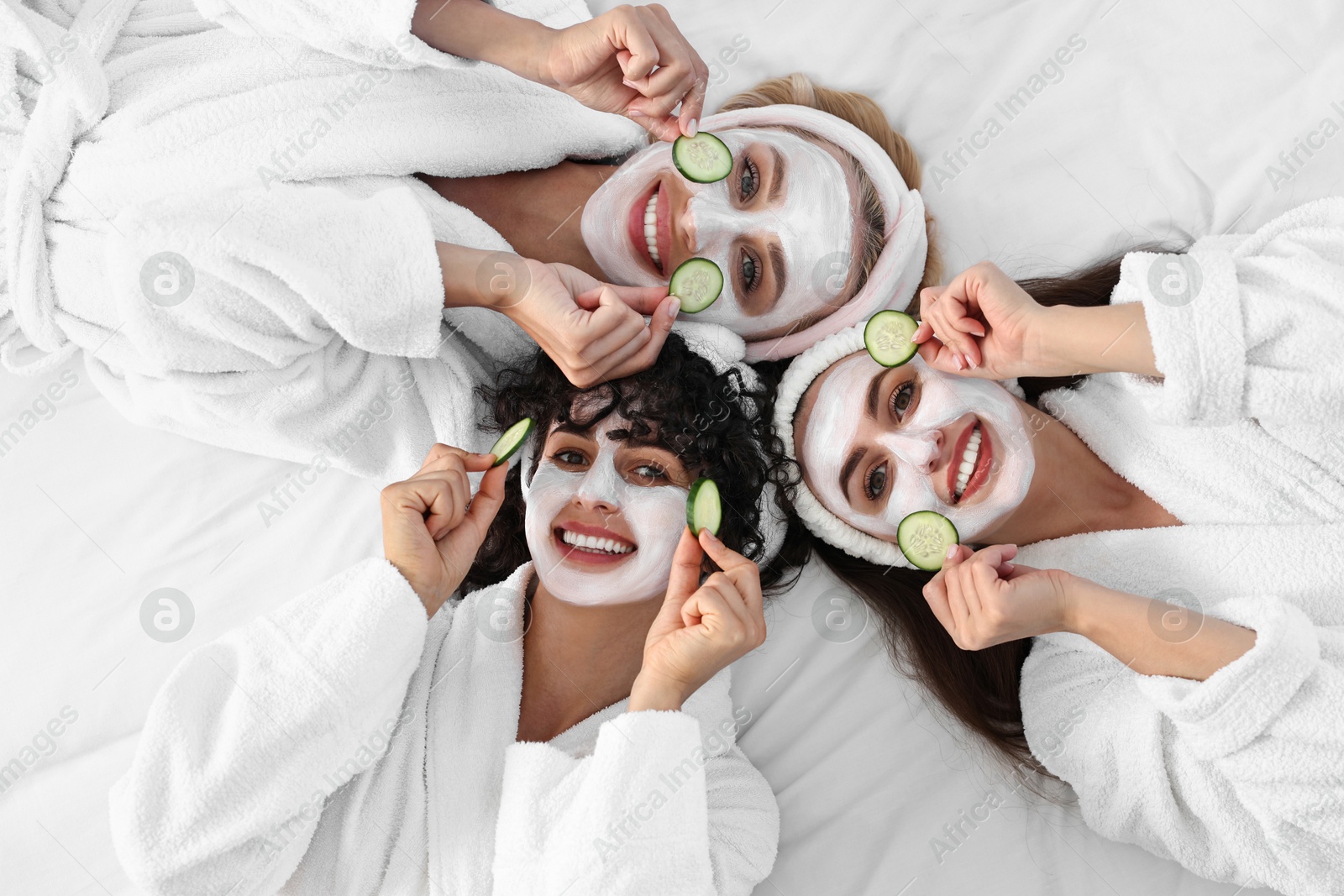 Photo of Happy friends with facial masks and cucumber slices on bed, top view. Spa party