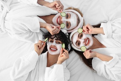 Photo of Happy friends with facial masks and cucumber slices on bed, top view. Spa party