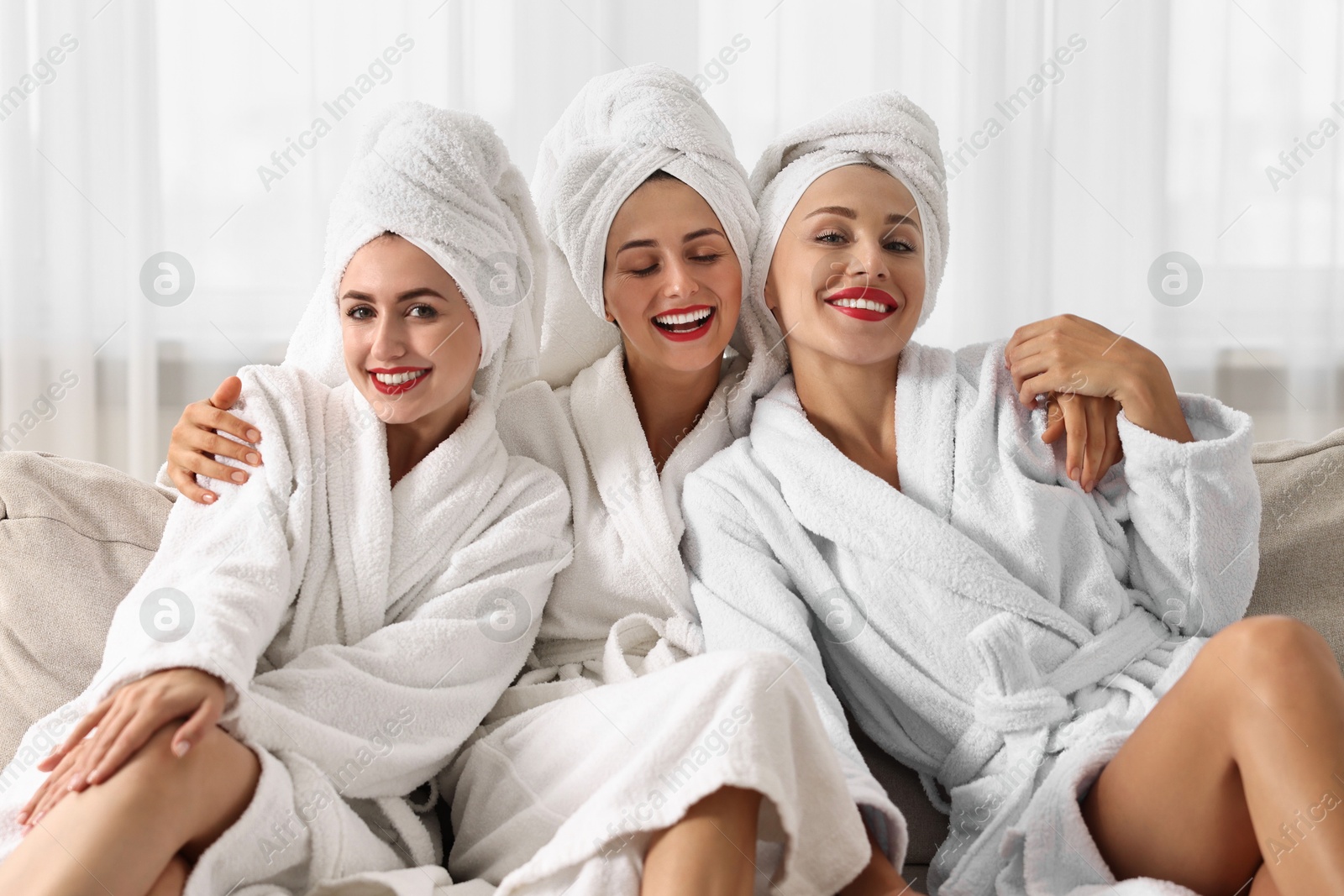 Photo of Happy friends in bathrobes with towels on couch indoors. Spa party