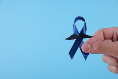 Prostate cancer awareness. Man holding blue ribbon with fake mustache on color background, closeup. Space for text