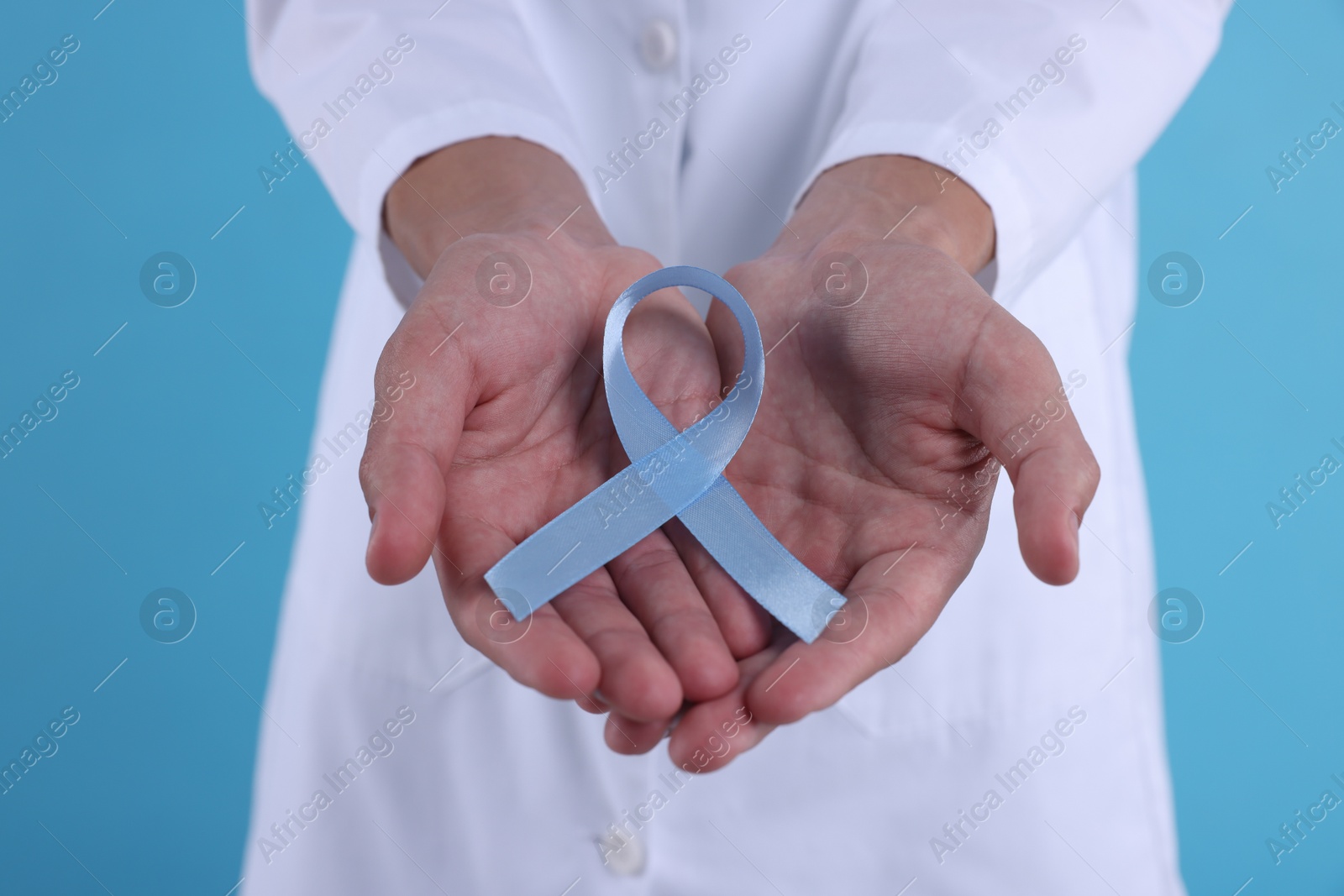 Photo of Prostate cancer awareness. Doctor holding light blue ribbon as symbol of support on color background, closeup