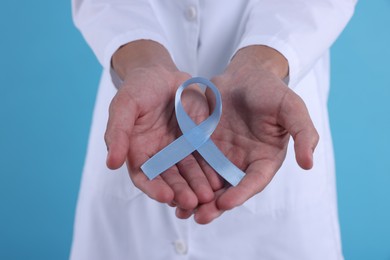 Photo of Prostate cancer awareness. Doctor holding light blue ribbon as symbol of support on color background, closeup