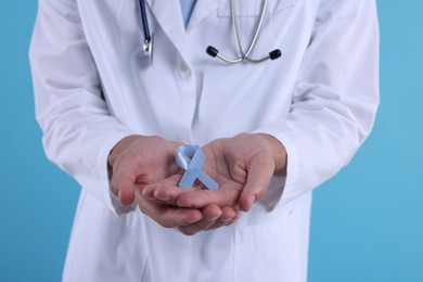 Photo of Prostate cancer awareness. Doctor holding light blue ribbon as symbol of support on color background, closeup