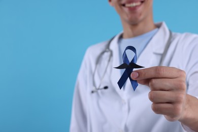Photo of Prostate cancer awareness. Doctor holding blue ribbon with fake mustache on color background, selective focus. Space for text