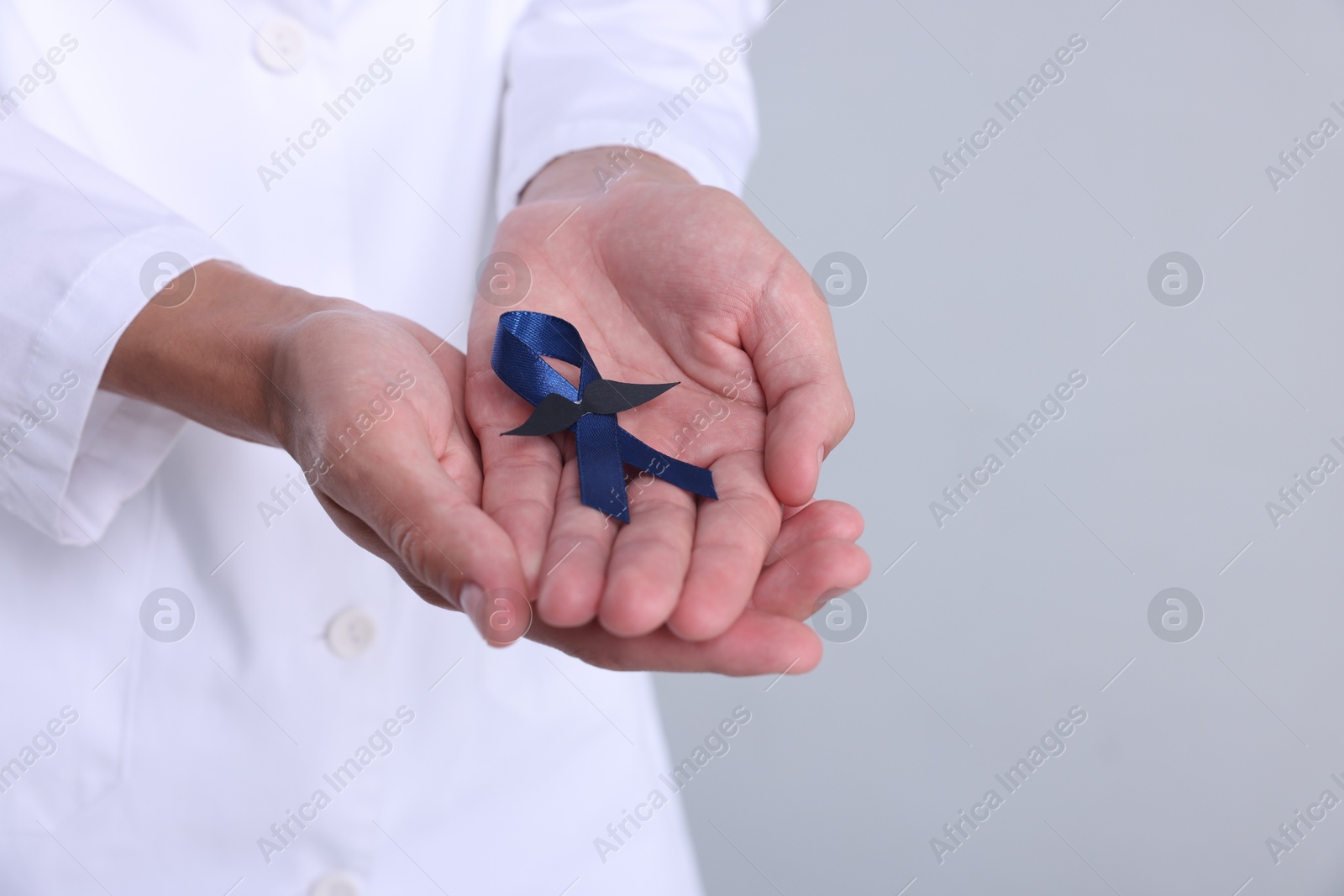 Photo of Prostate cancer awareness. Doctor holding blue ribbon with fake mustache on light grey background, closeup. Space for text