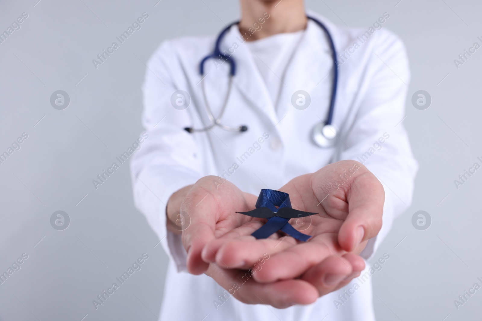 Photo of Prostate cancer awareness. Doctor holding blue ribbon with fake mustache on light grey background, closeup