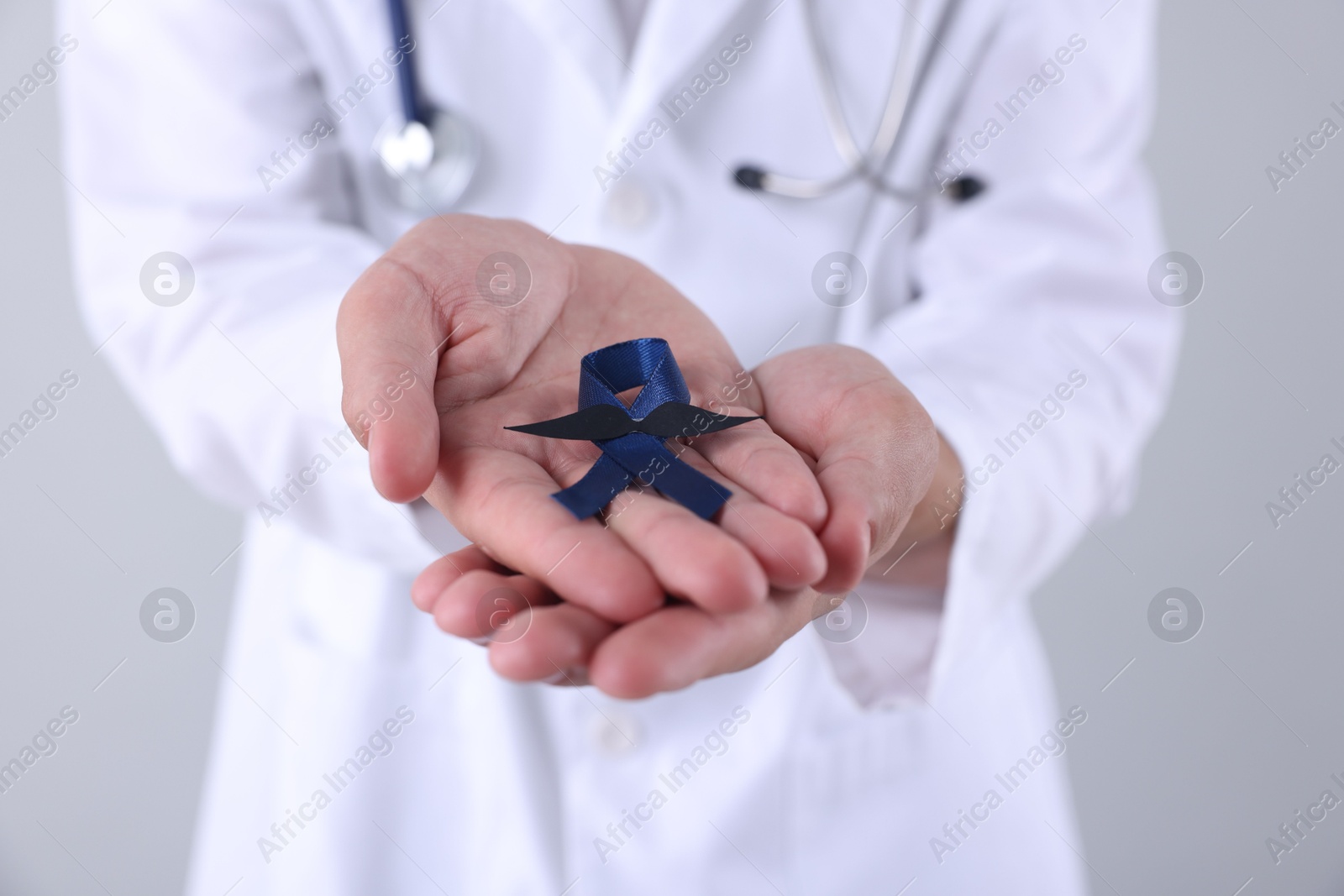 Photo of Prostate cancer awareness. Doctor holding blue ribbon with fake mustache on light grey background, closeup