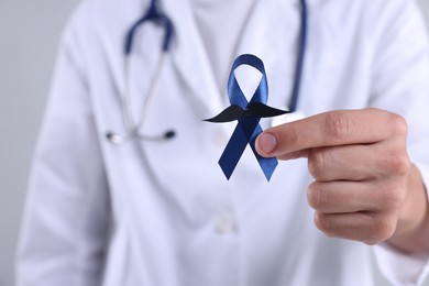 Photo of Prostate cancer awareness. Doctor holding blue ribbon with fake mustache on light grey background, closeup