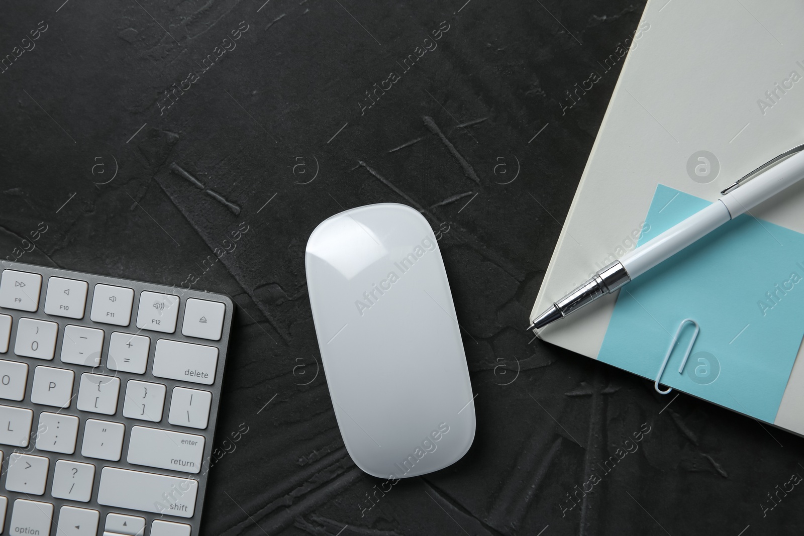 Photo of Wireless mouse, computer keyboard, notebook and pen on dark textured table, flat lay