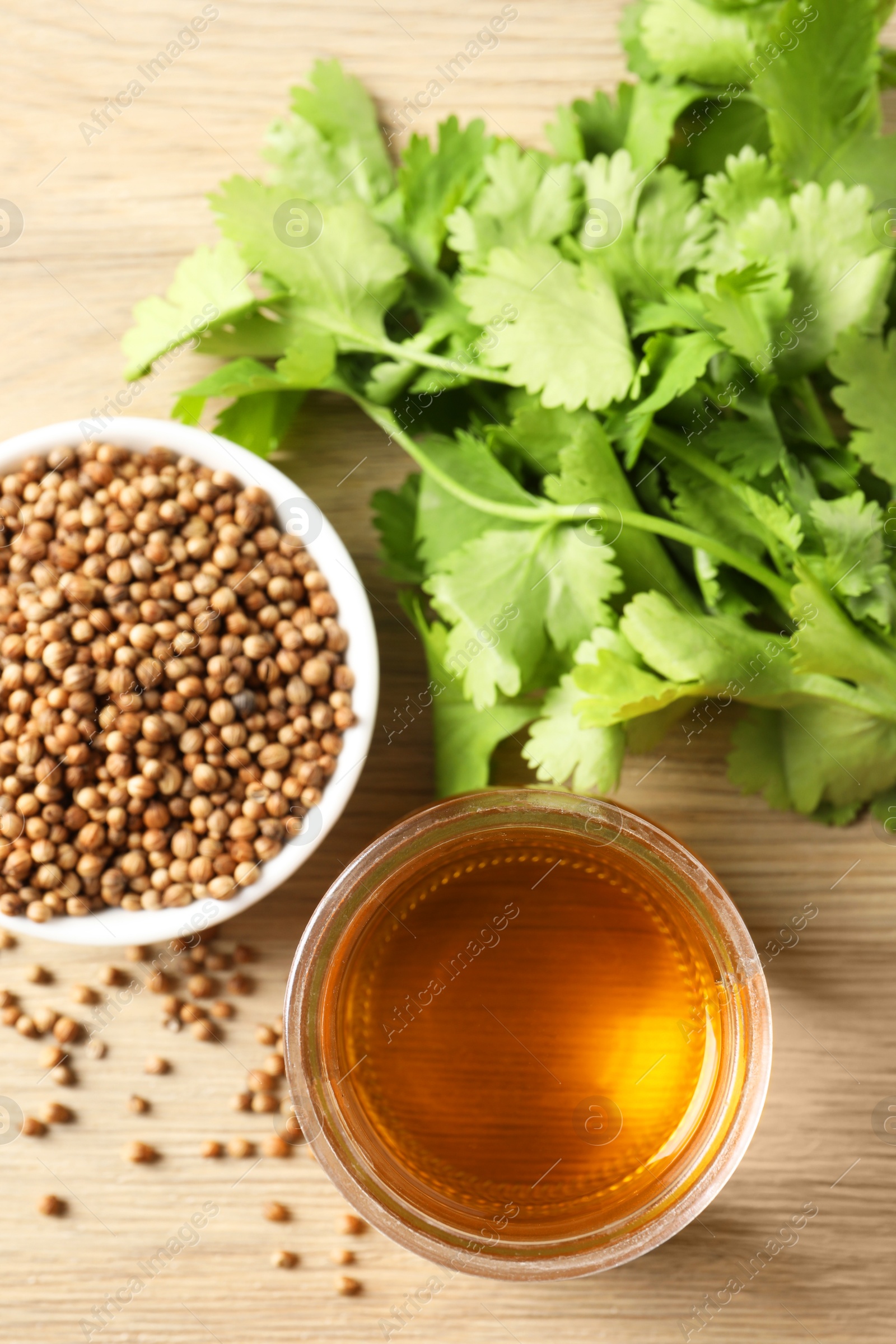 Photo of Dried coriander seeds in bowl, oil and green leaves on wooden table, top view