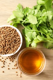 Dried coriander seeds in bowl, oil and green leaves on wooden table, top view