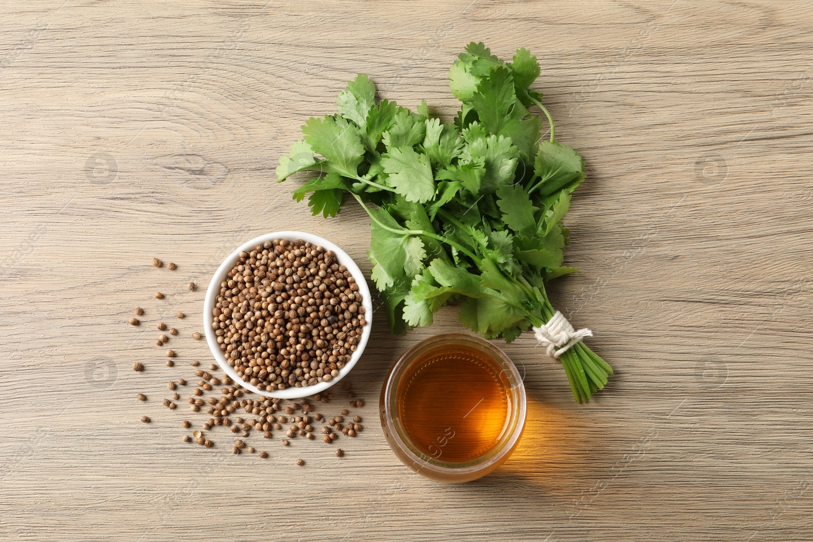 Photo of Dried coriander seeds in bowl, oil and green leaves on wooden table, top view