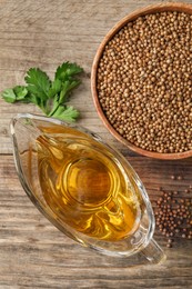 Photo of Dried coriander seeds in bowl, oil and green leaves on wooden table, top view