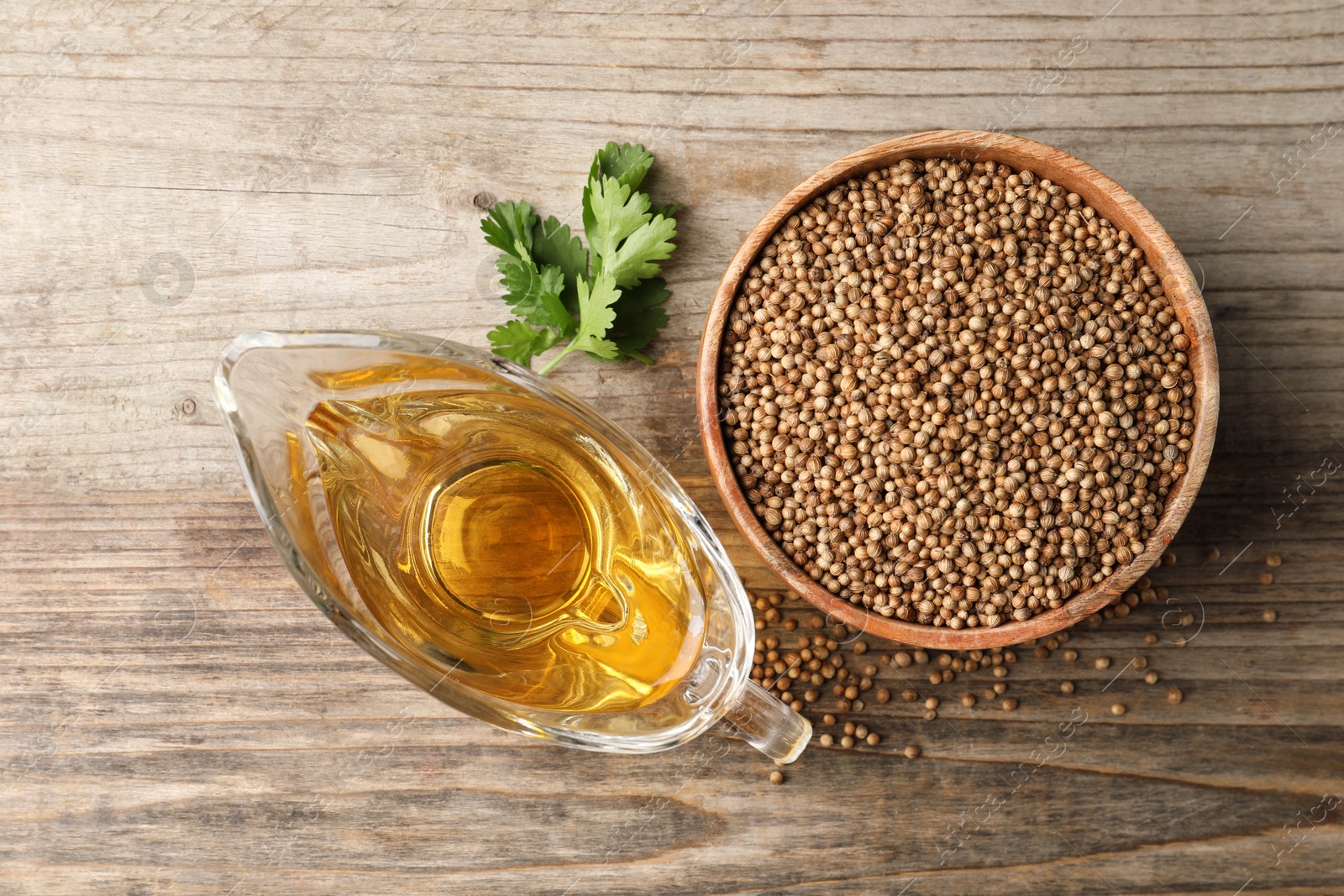 Photo of Dried coriander seeds in bowl, oil and green leaves on wooden table, top view