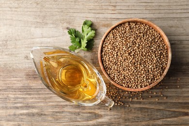 Photo of Dried coriander seeds in bowl, oil and green leaves on wooden table, top view