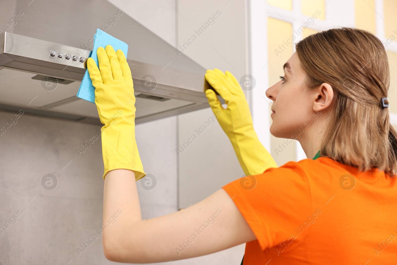 Photo of Professional janitor wearing uniform cleaning kitchen hood with rag indoors