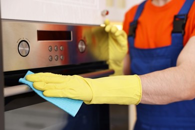 Photo of Professional janitor wearing uniform cleaning electric oven in kitchen, closeup