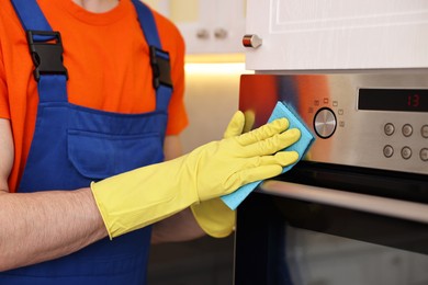 Photo of Professional janitor wearing uniform cleaning electric oven in kitchen, closeup