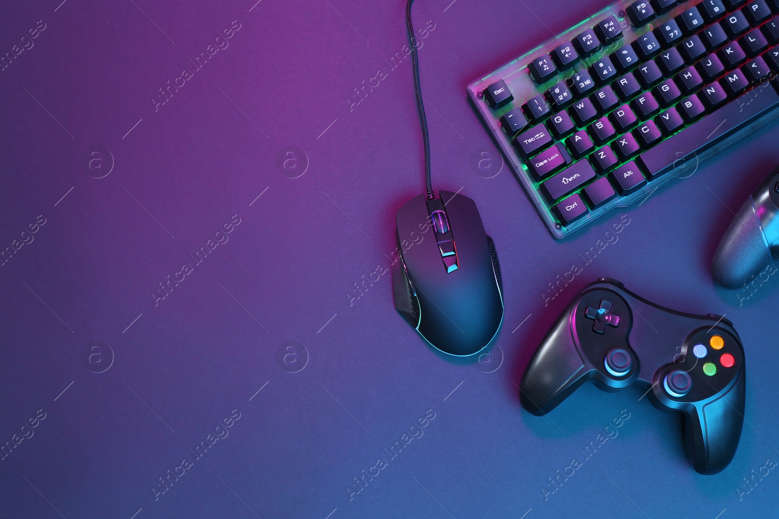 Photo of Computer mouse, RGB keyboard and game controllers in neon lights on dark background, flat lay. Space for text