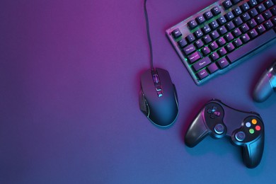 Photo of Computer mouse, RGB keyboard and game controllers in neon lights on dark background, flat lay. Space for text