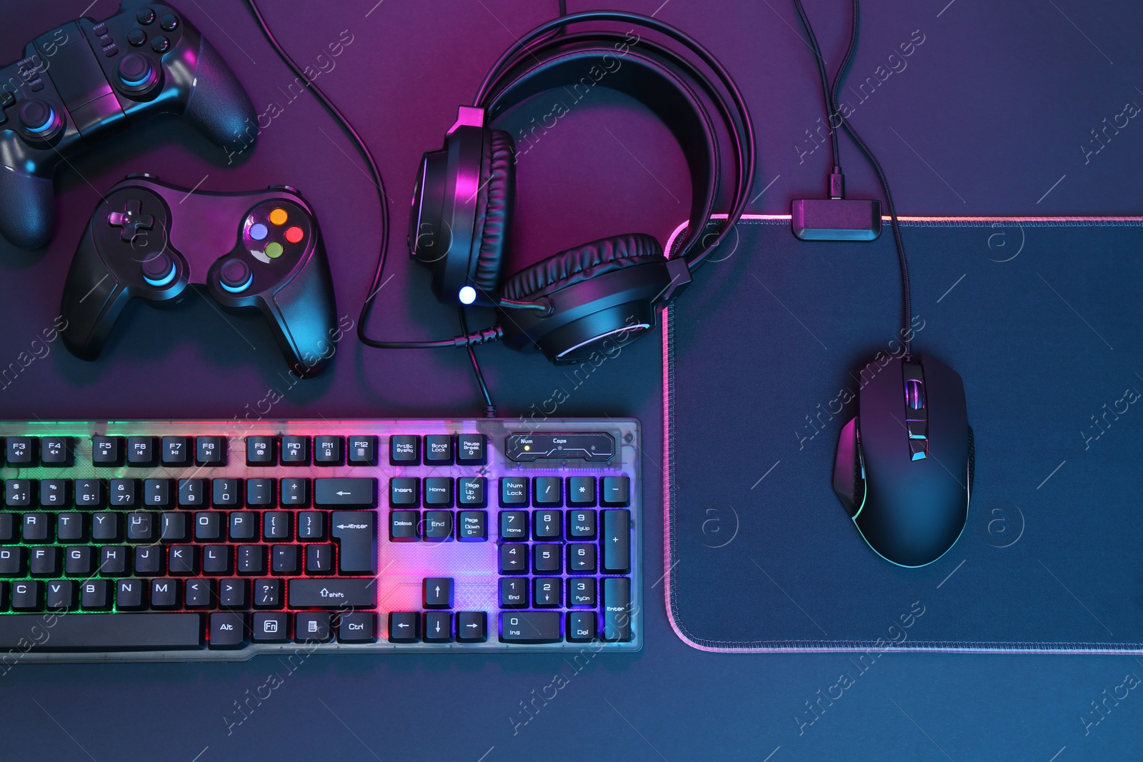 Photo of Computer mouse, RGB keyboard, headset and game controllers in neon lights on dark background, top view