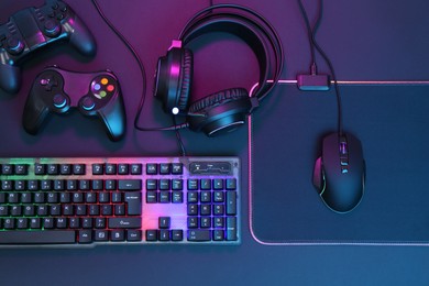 Computer mouse, RGB keyboard, headset and game controllers in neon lights on dark background, top view