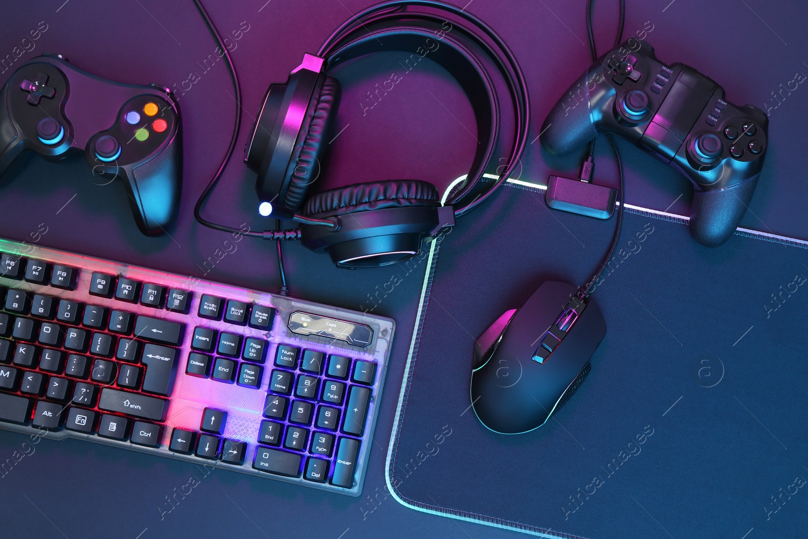 Photo of Computer mouse, RGB keyboard, headset and game controllers in neon lights on dark background, top view