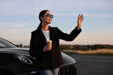 Photo of Smiling woman in sunglasses with paper cup of drink near car outdoors